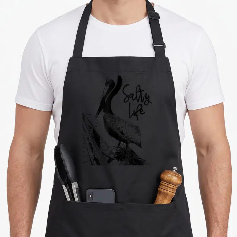 Salty pelican apron - Black line art pelican on pier with stacked Salty Life text — shown on a black cotton canvas apron against a white background