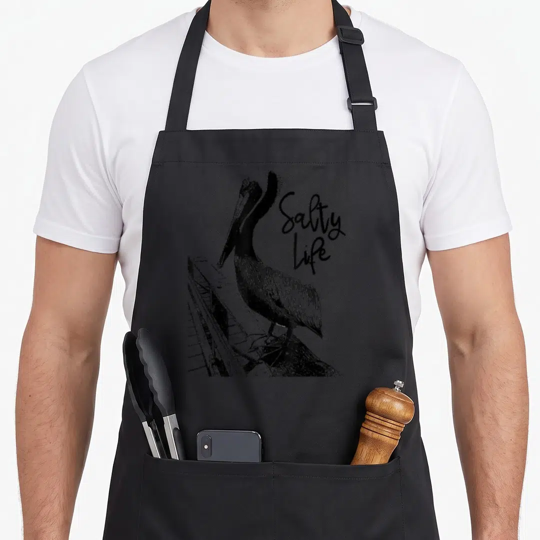 Salty pelican apron - Black line art pelican on pier with stacked Salty Life text — shown on a black cotton canvas apron against a white background