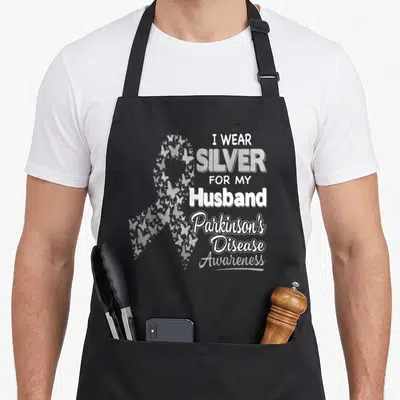 Parkinson husband apron - Silver ribbon formed by butterflies next to stacked text about wearing silver for a husband — shown on a black cotton canvas apron against a white background