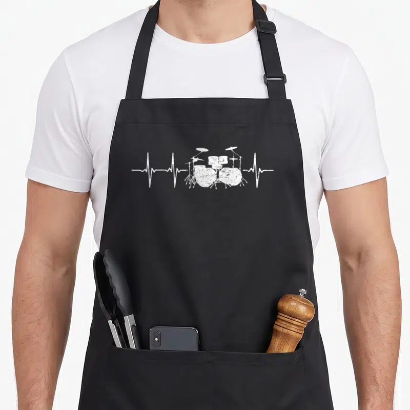 Drum beat apron - White distressed drum kit with horizontal heartbeat line extending from both sides — shown on a black cotton canvas apron against a white background