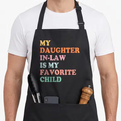 Funny law mother daughter apron - Stacked text reading MY DAUGHTER IN-LAW IS MY FAVORITE CHILD in yellow, orange, pink — shown on a black cotton canvas apron against a white background