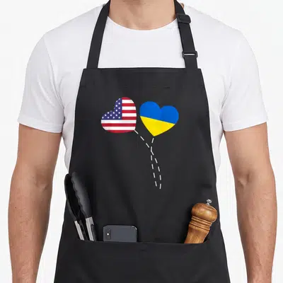 Loving american flag apron - Two heart-shaped US and Ukraine flags connected by white dashed lines in red white blue — shown on a black cotton canvas apron against a white background