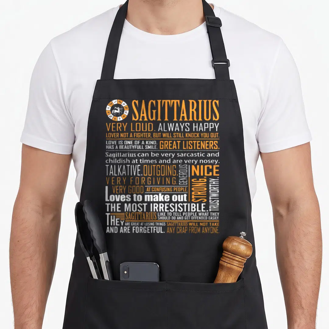 Loud zodiac aprons - Orange and white Sagittarius personality text block with zodiac wheel icon — shown on a black cotton canvas apron against a white background