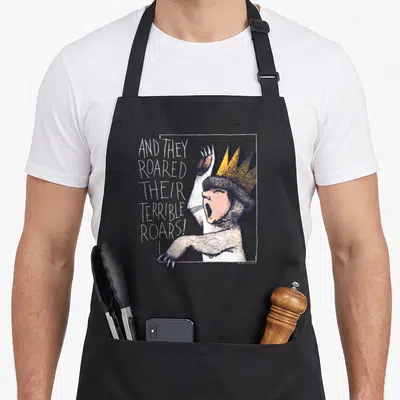 Roar wild apron - Crowned grey and white cat roaring next to stacked white text inside a white frame — shown on a black cotton canvas apron against a white background