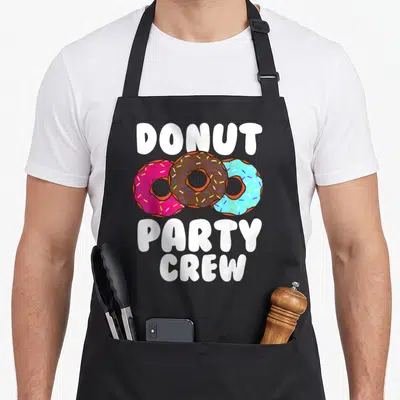 Doughnut crew apron - Three cartoon donuts in pink brown and blue with white text reading DONUT PARTY CREW — shown on a black cotton canvas apron against a white background