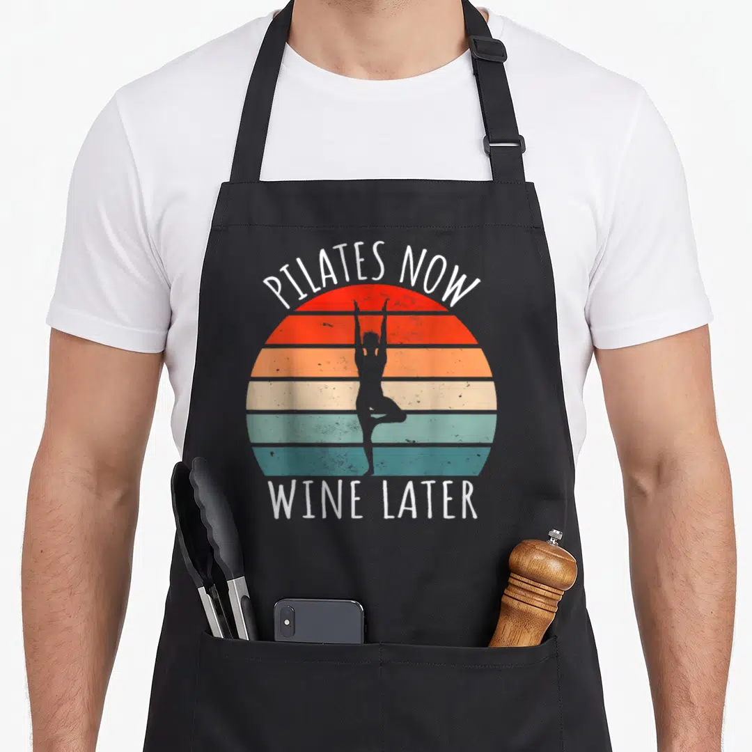Meditation pilates apron - Retro striped sun with Pilates silhouette and white text PILATES NOW WINE LATER — shown on a black cotton canvas apron against a white background