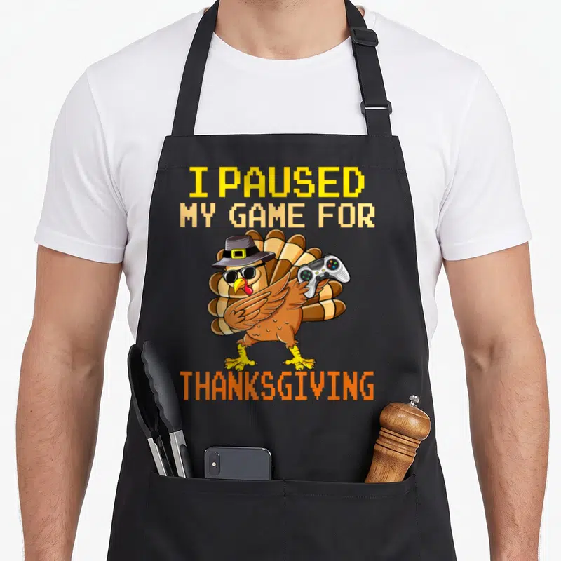 Gaming fall thanksgiving apron - Cartoon turkey in pilgrim hat holding controller under pixel text I PAUSED MY GAME — shown on a black cotton canvas apron against a white background