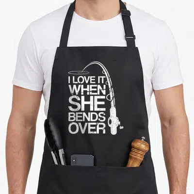 Funny she fishing aprons - Distressed white fishing rod curving over stacked text I LOVE IT WHEN SHE BENDS — shown on a black cotton canvas apron against a white background