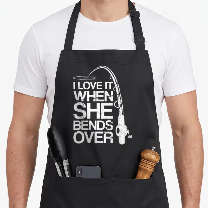 Funny she fishing aprons - Distressed white fishing rod curving over stacked text I LOVE IT WHEN SHE BENDS — shown on a black cotton canvas apron against a white background
