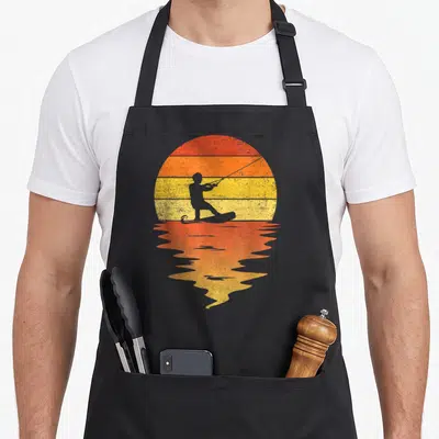 Wakeboarding 70s apron - Retro wakeboarder silhouette on striped red orange yellow sun with water reflection — shown on a black cotton canvas apron against a white background