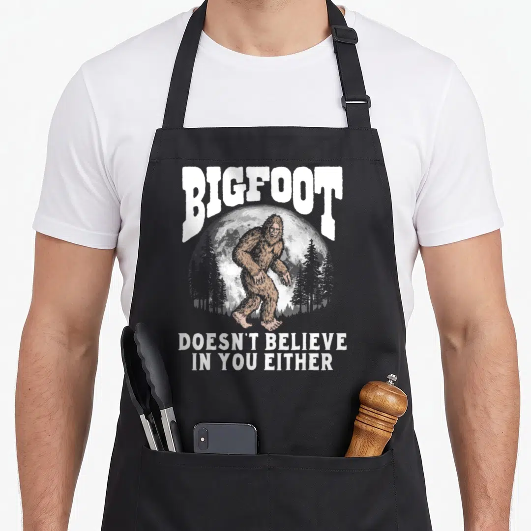 Moon bigfoot apron - Brown Bigfoot figure in front of white moon with arched text and stacked slogan below — shown on a black cotton canvas apron against a white background