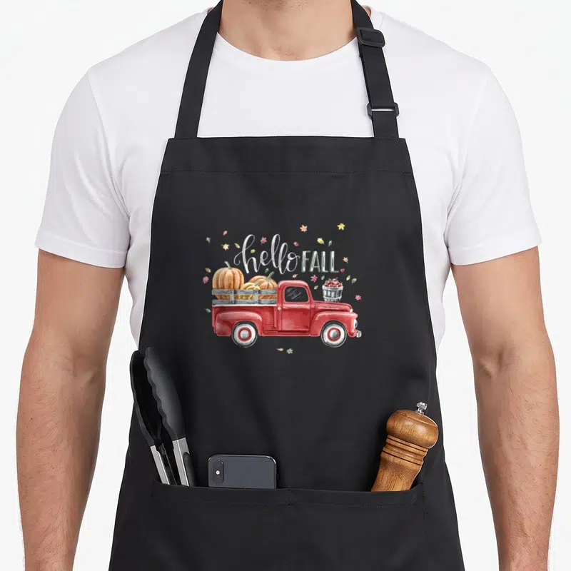 Hello fall apron - Red truck carrying two orange pumpkins and a bucket of red apples under hello FALL text — shown on a black cotton canvas apron against a white background