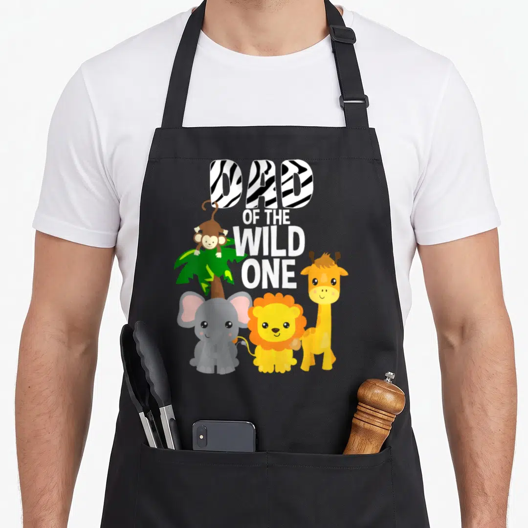 Jungle birthday apron - DAD OF THE WILD ONE text with cartoon monkey, elephant, lion, giraffe, and zebra stripe letters — shown on a black cotton canvas apron against a white background