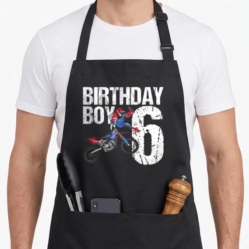 Bike birthday apron - Cartoon motocross rider jumping in front of white BIRTHDAY BOY 6 text in red blue and black — shown on a black cotton canvas apron against a white background