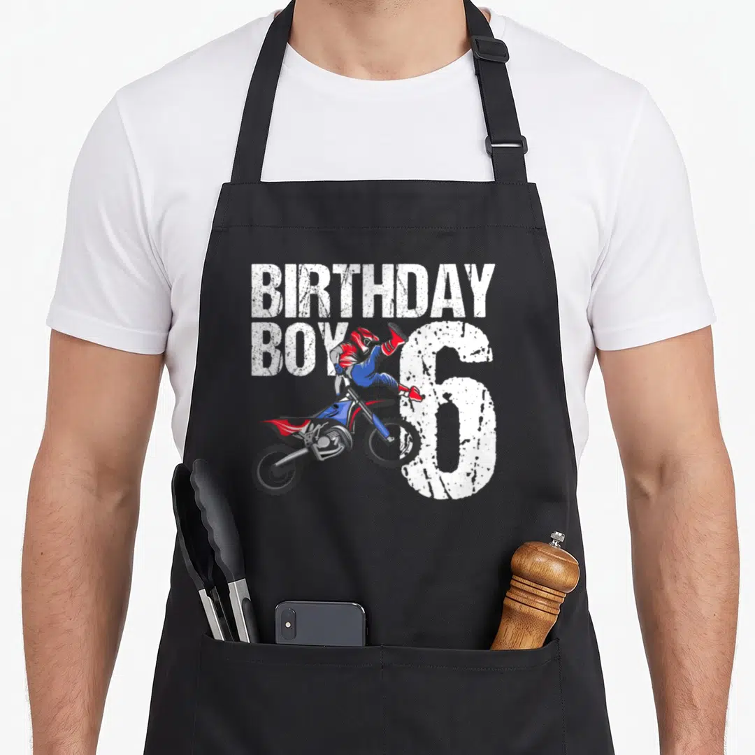 Bike birthday apron - Cartoon motocross rider jumping in front of white BIRTHDAY BOY 6 text in red blue and black — shown on a black cotton canvas apron against a white background