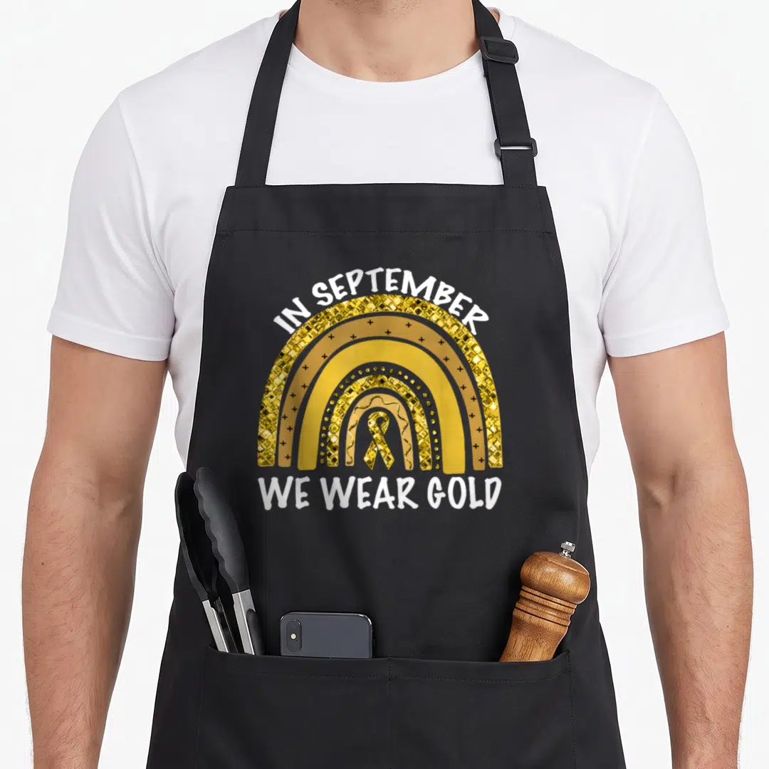 September childhood cancer awareness apron - Gold retro rainbow with white text IN SEPTEMBER WE WEAR GOLD and central — shown on a black cotton canvas apron against a white background
