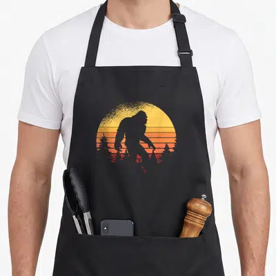 Believer bigfoot apron - Black Bigfoot silhouette walking past a striped yellow orange and red retro sun with trees — shown on a black cotton canvas apron against a white background