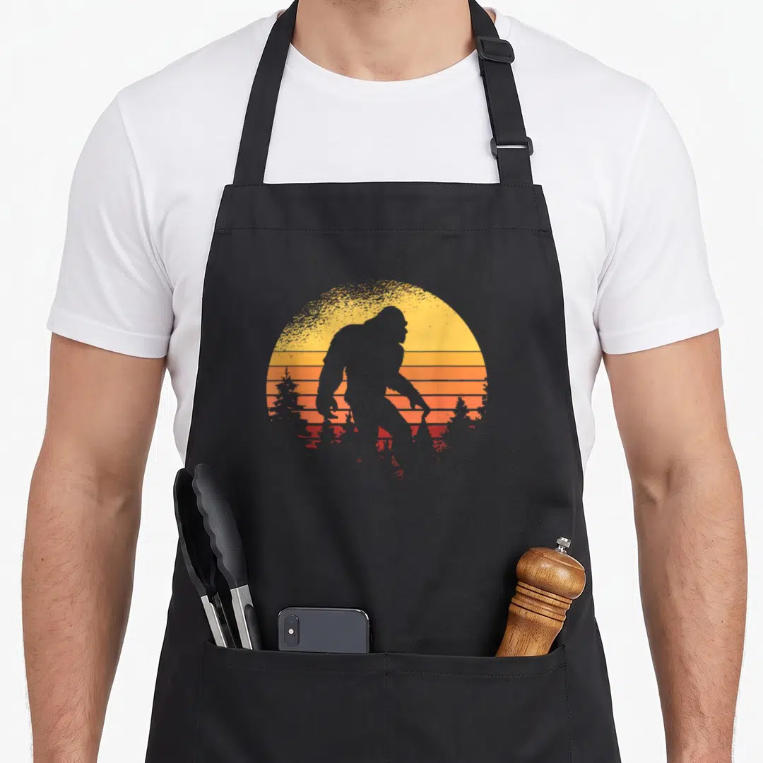 Believer bigfoot apron - Black Bigfoot silhouette walking past a striped yellow orange and red retro sun with trees — shown on a black cotton canvas apron against a white background