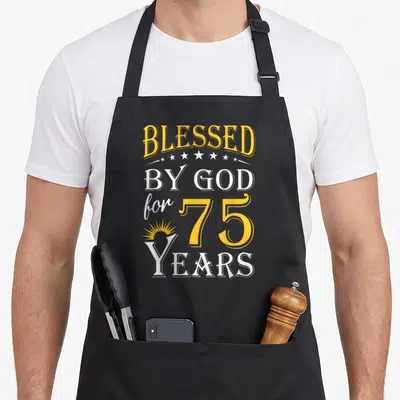 God 75th birthday apron - Vertical yellow and white text BLESSED BY GOD for 75 YEARS with stars and sunburst — shown on a black cotton canvas apron against a white background