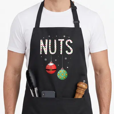 Funny nut couples christmas aprons - NUTS text with red Santa ornament and green star ornament hanging below — shown on a black cotton canvas apron against a white background