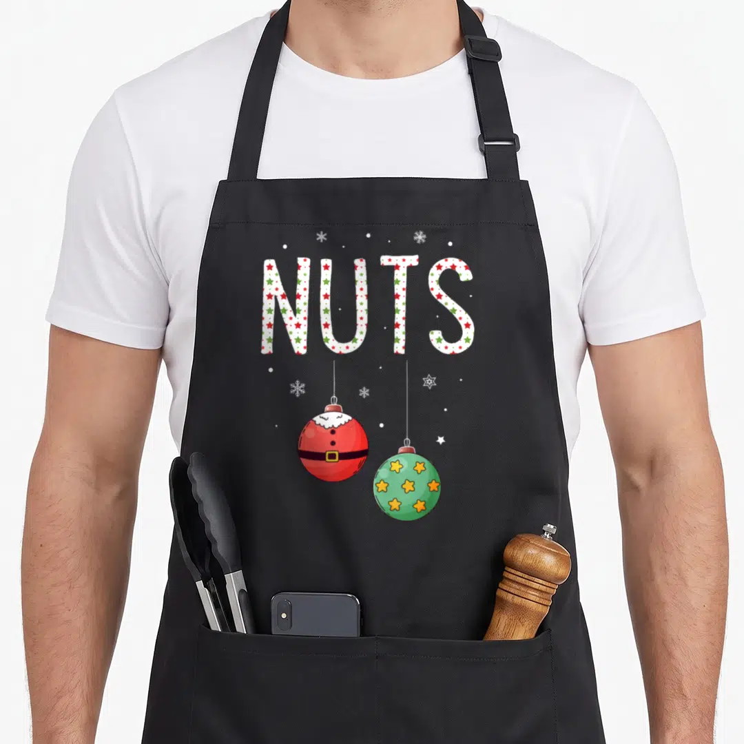 Funny nut couples christmas aprons - NUTS text with red Santa ornament and green star ornament hanging below — shown on a black cotton canvas apron against a white background