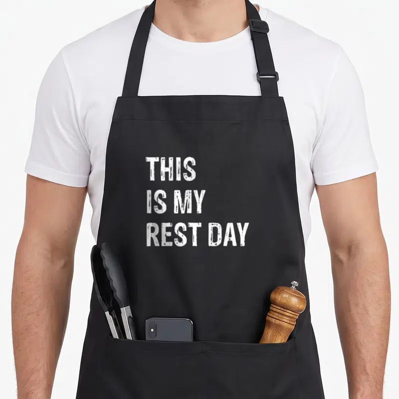 Rest sarcastic apron - White stacked text reading THIS IS MY REST DAY on dark background — shown on a black cotton canvas apron against a white background