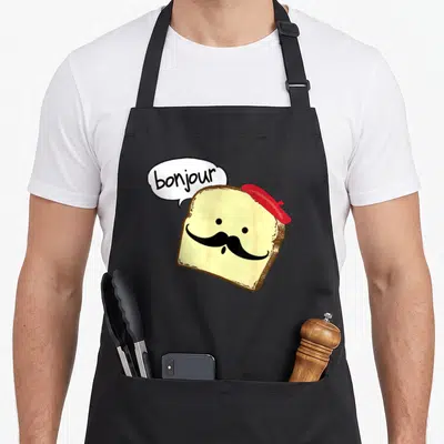 Bonjour toast apron - Cartoon bread slice with red beret, black mustache, and speech bubble reading bonjour — shown on a black cotton canvas apron against a white background