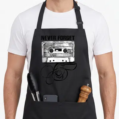 Music cassette apron - Black and white line art of a cassette tape with spilling ribbon and NEVER FORGET text — shown on a black cotton canvas apron against a white background