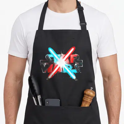 Dueling star apron - Two crossed lightsabers over STAR WARS text in blue red silver white — shown on a black cotton canvas apron against a white background
