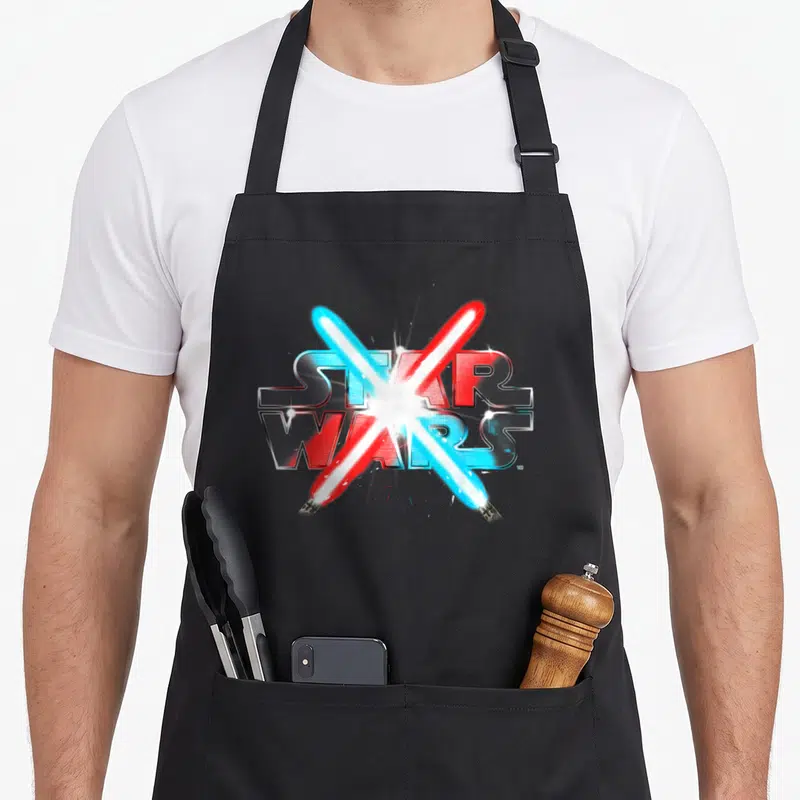 Dueling star apron - Two crossed lightsabers over STAR WARS text in blue red silver white — shown on a black cotton canvas apron against a white background
