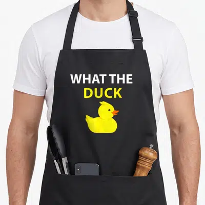 Funny duck rubber apron - Stacked white and yellow text WHAT THE DUCK above a yellow cartoon rubber duck — shown on a black cotton canvas apron against a white background
