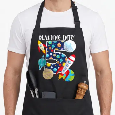 Blasting birthday apron - BLASTING INTO text above space-patterned number 5 flanked by two rockets and planets — shown on a black cotton canvas apron against a white background