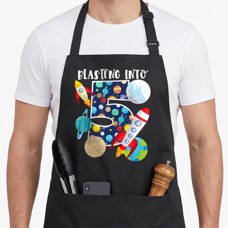Blasting birthday apron - BLASTING INTO text above space-patterned number 5 flanked by two rockets and planets — shown on a black cotton canvas apron against a white background