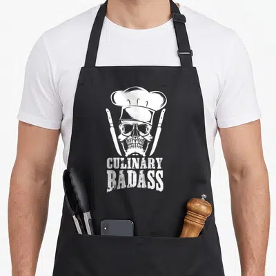 Badass culinary aprons - White skull in chef hat flanked by two knives with stacked text — shown on a black cotton canvas apron against a white background