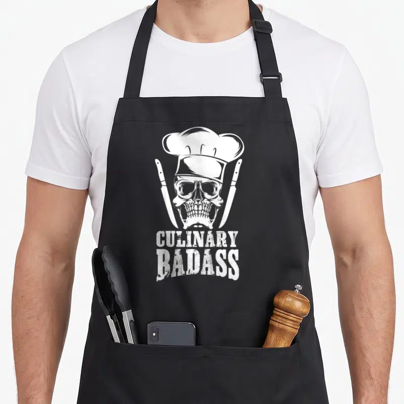 Badass culinary aprons - White skull in chef hat flanked by two knives with stacked text — shown on a black cotton canvas apron against a white background