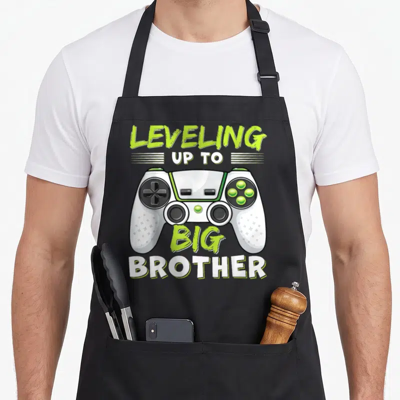 Leveling big brother apron - Game controller in white and lime green with LEVELING UP TO BIG BROTHER text in matching — shown on a black cotton canvas apron against a white background