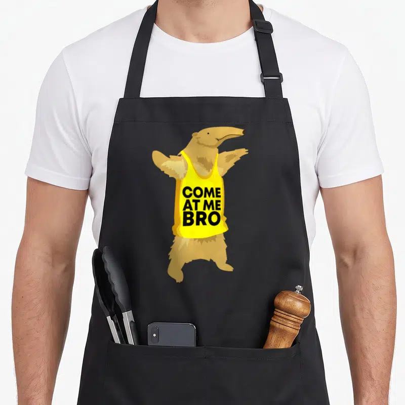 Come cartoon apron - Tan anteater in yellow tank top with black COME AT ME BRO text — shown on a black cotton canvas apron against a white background