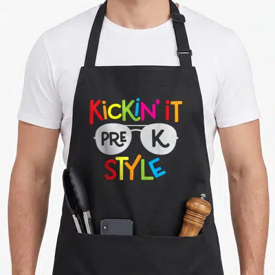 Kickin teacher apron - Cartoon glasses with PRE K inside lenses and Kickin It Style text in rainbow colors — shown on a black cotton canvas apron against a white background