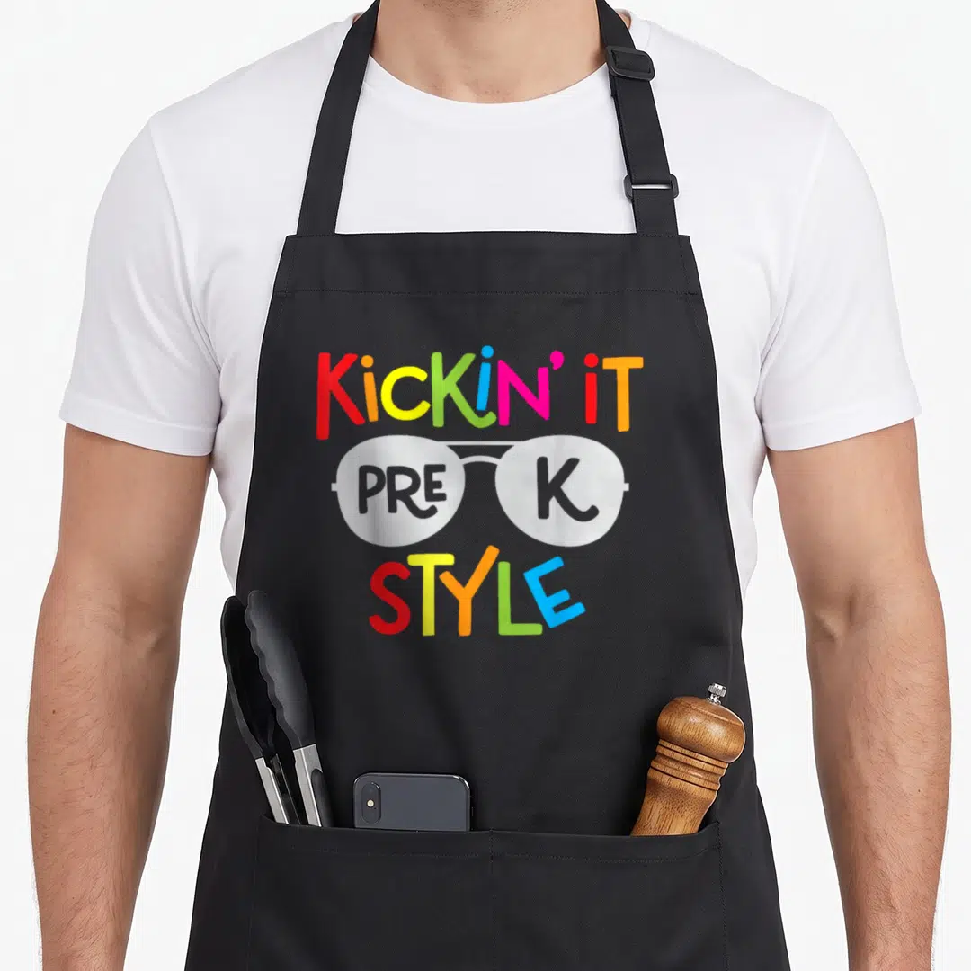 Kickin teacher apron - Cartoon glasses with PRE K inside lenses and Kickin It Style text in rainbow colors — shown on a black cotton canvas apron against a white background