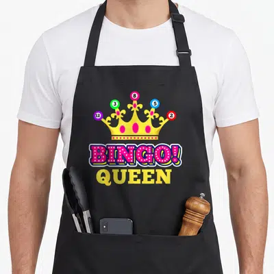 Queen bingo apron - Yellow crown with numbered balls and pink BINGO! QUEEN text — shown on a black cotton canvas apron against a white background