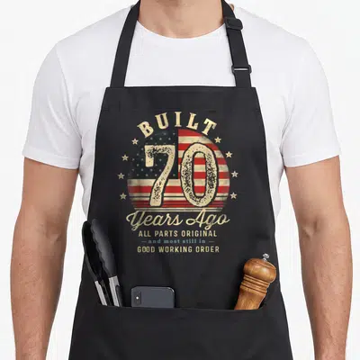 Ago 70th birthday apron - BUILT 70 Years Ago text over distressed circular USA flag with red white and blue stars — shown on a black cotton canvas apron against a white background