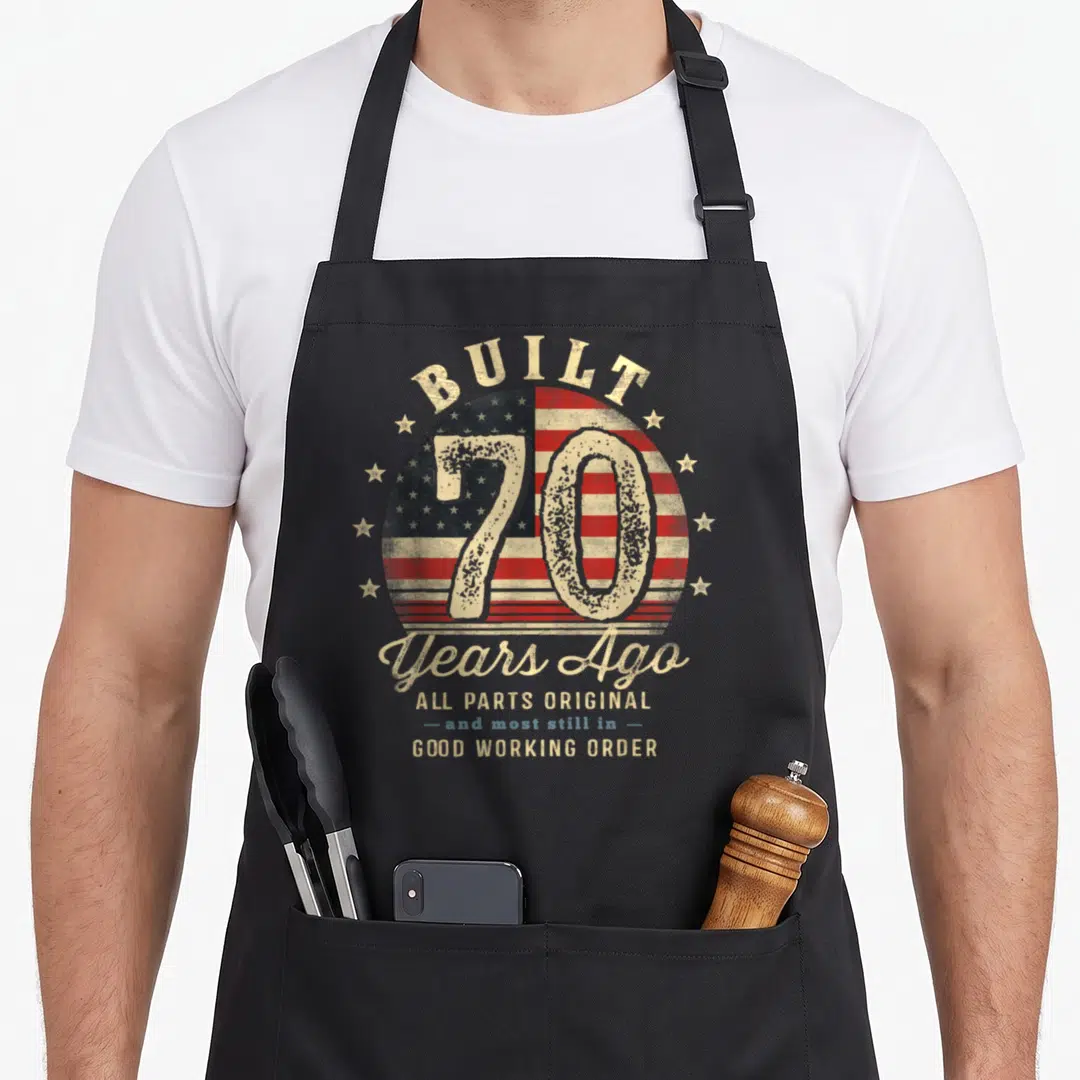 Ago 70th birthday apron - BUILT 70 Years Ago text over distressed circular USA flag with red white and blue stars — shown on a black cotton canvas apron against a white background