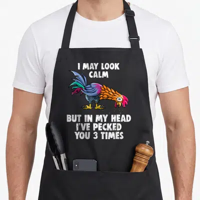 But pecked you apron - Cartoon rooster in blue purple orange and red surrounded by white text reading I may look calm — shown on a black cotton canvas apron against a white background