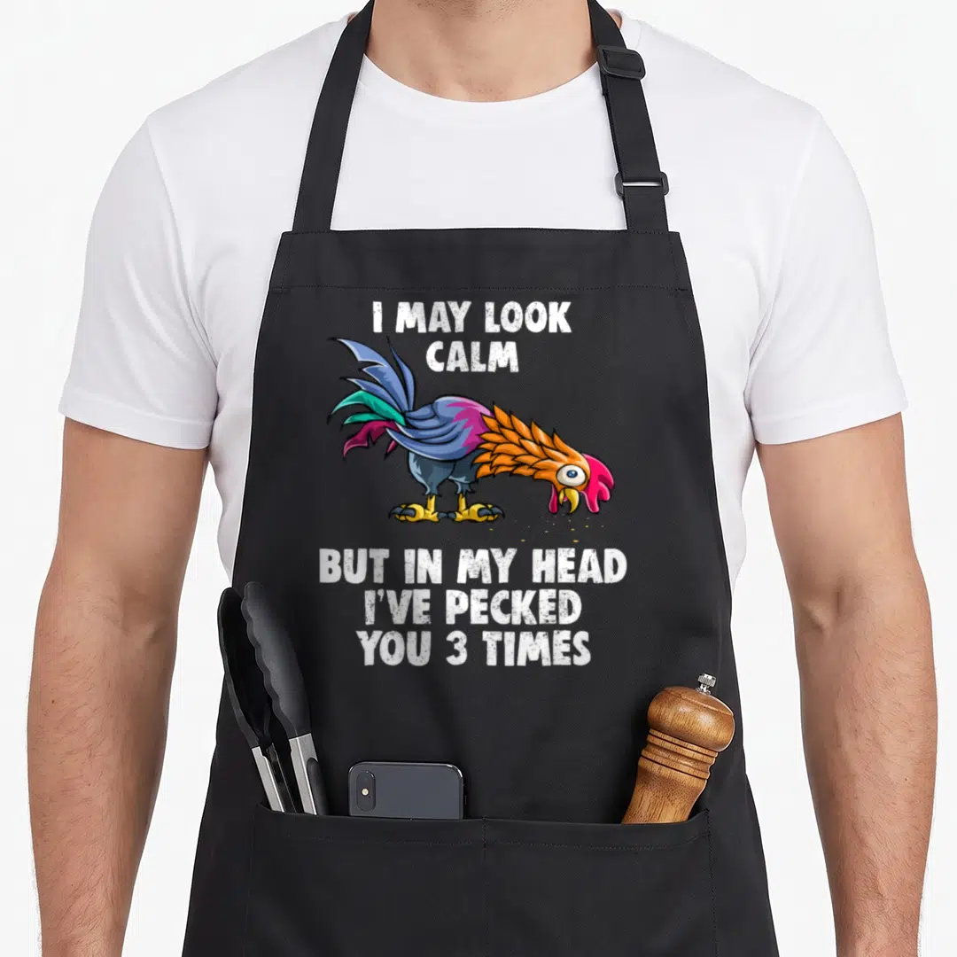 But pecked you apron - Cartoon rooster in blue purple orange and red surrounded by white text reading I may look calm — shown on a black cotton canvas apron against a white background