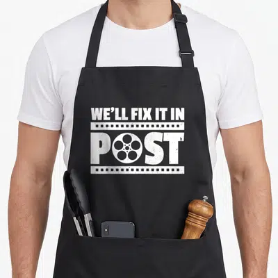 Post filmmaking apron - White distressed We'll Fix It In Post text with film strip and reel graphic — shown on a black cotton canvas apron against a white background