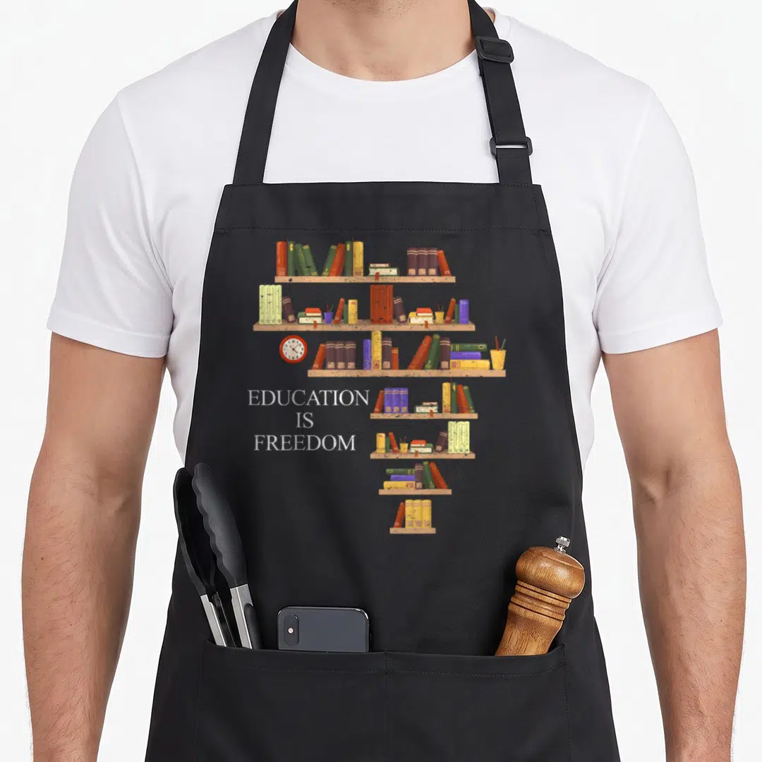 Month history apron - Stacked text education is freedom next to illustrated bookshelves with colorful books and a clock — shown on a black cotton canvas apron against a white background