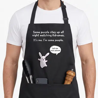 Army drama apron - Pink cartoon bunny with speech bubble next to white text about staying up all night watching Kdramas — shown on a black cotton canvas apron against a white background