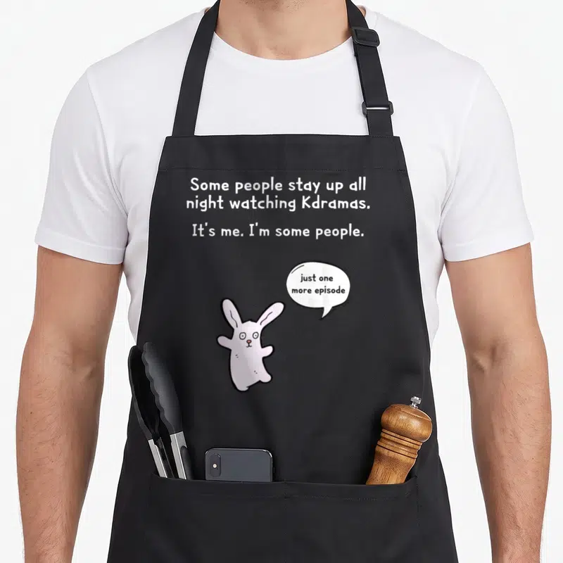 Army drama apron - Pink cartoon bunny with speech bubble next to white text about staying up all night watching Kdramas — shown on a black cotton canvas apron against a white background