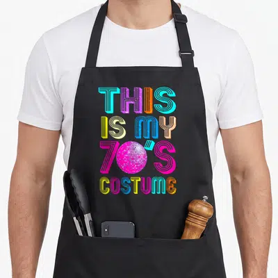 Music halloween apron - Retro text THIS IS MY 70S COSTUME with pink disco ball replacing the zero in colorful letters — shown on a black cotton canvas apron against a white background