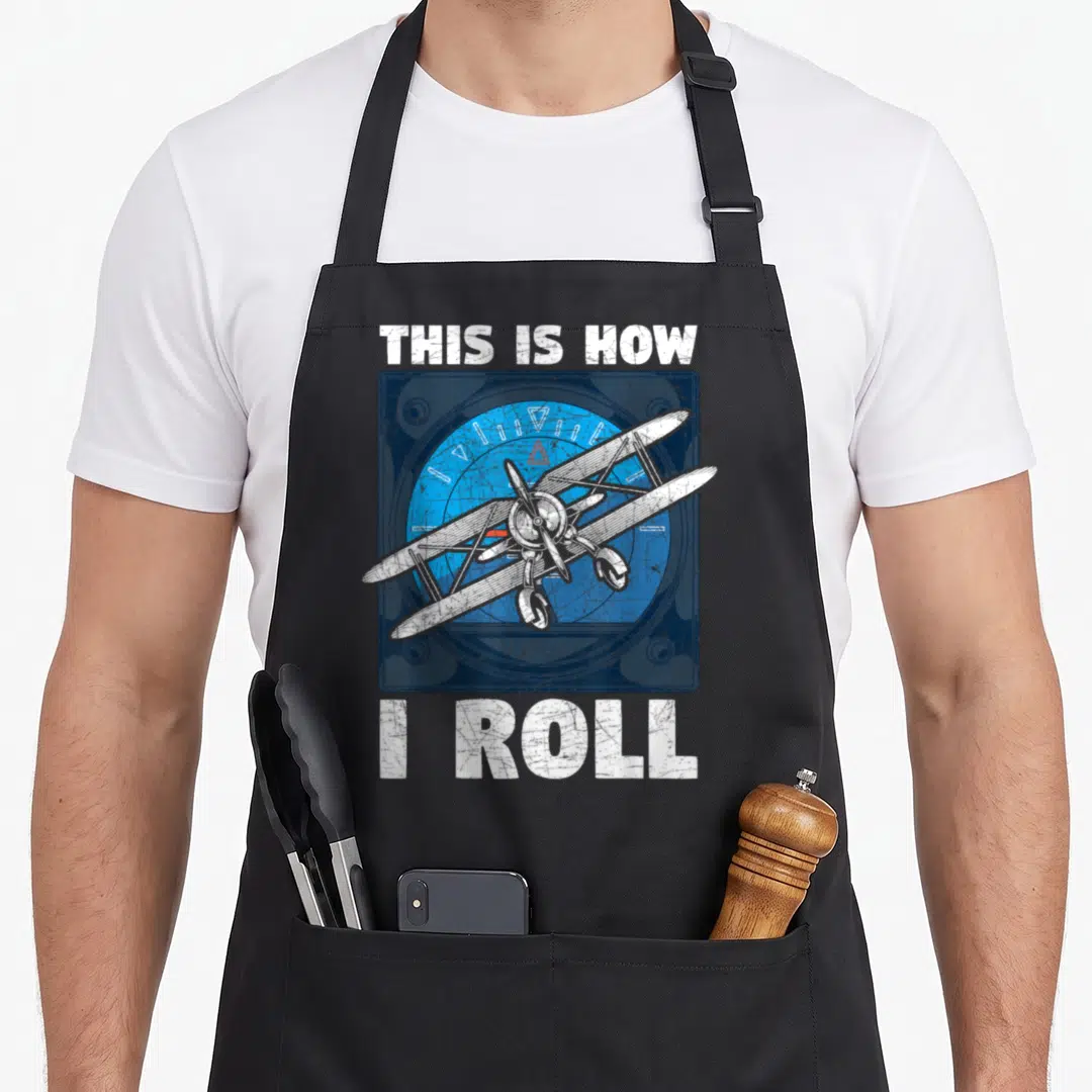 Bin nun apron - White biplane over blue circular dial with text This Is How I Roll — shown on a black cotton canvas apron against a white background